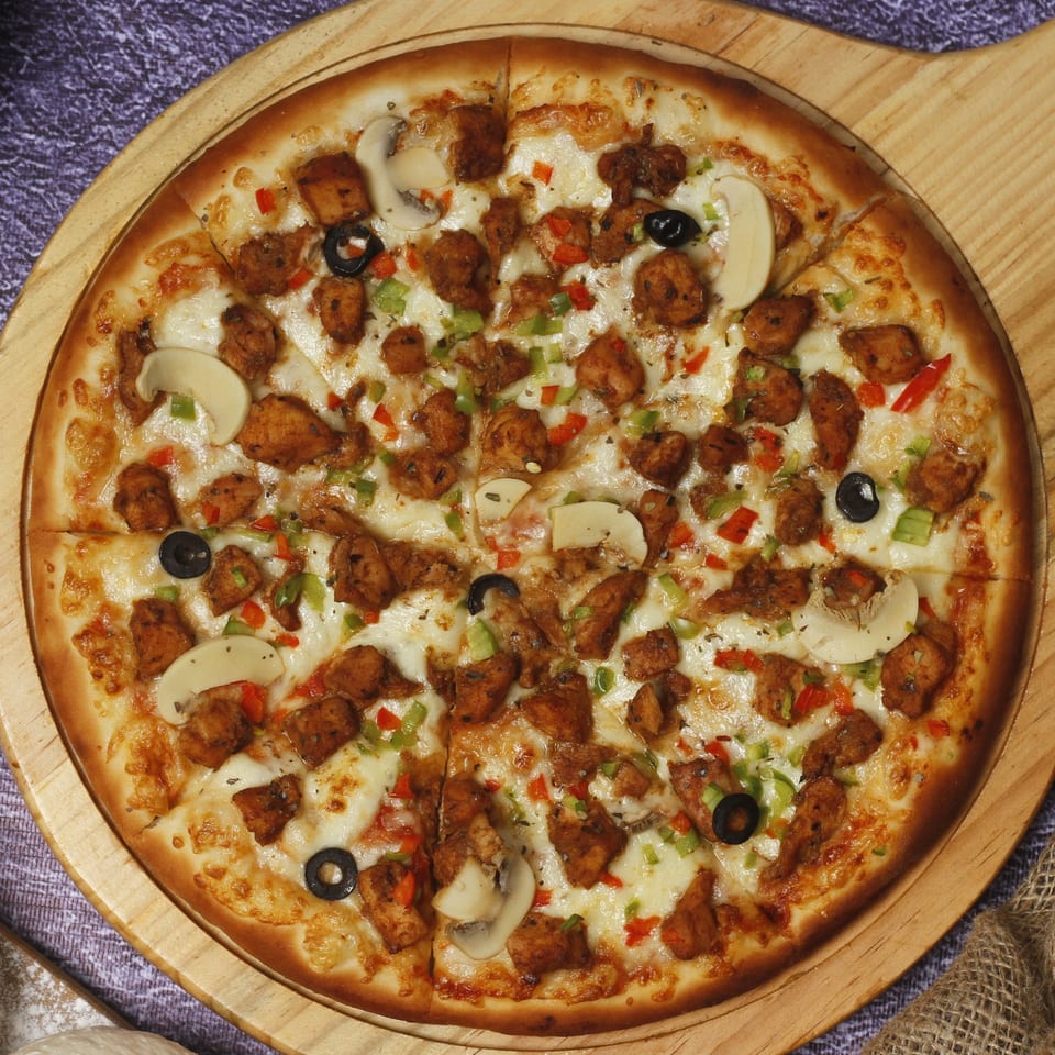 A Vincents pizza with chicken, mushrooms, black olives, and bell peppers on a wooden board.
