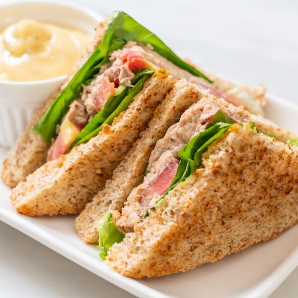Four triangular tuna salad sandwiches with lettuce and tomato, served with a side of sauce.