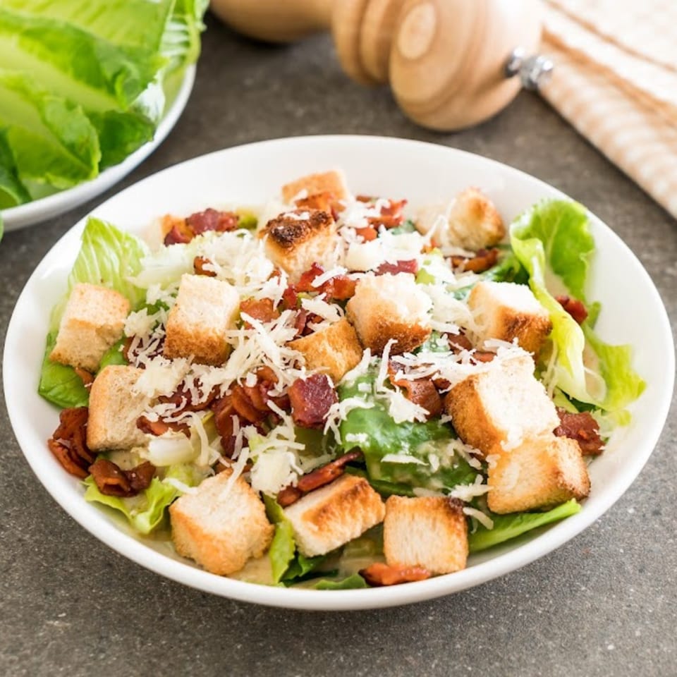 Caesar salad with romaine lettuce, croutons, bacon bits, and shredded Parmesan cheese in a white bowl.